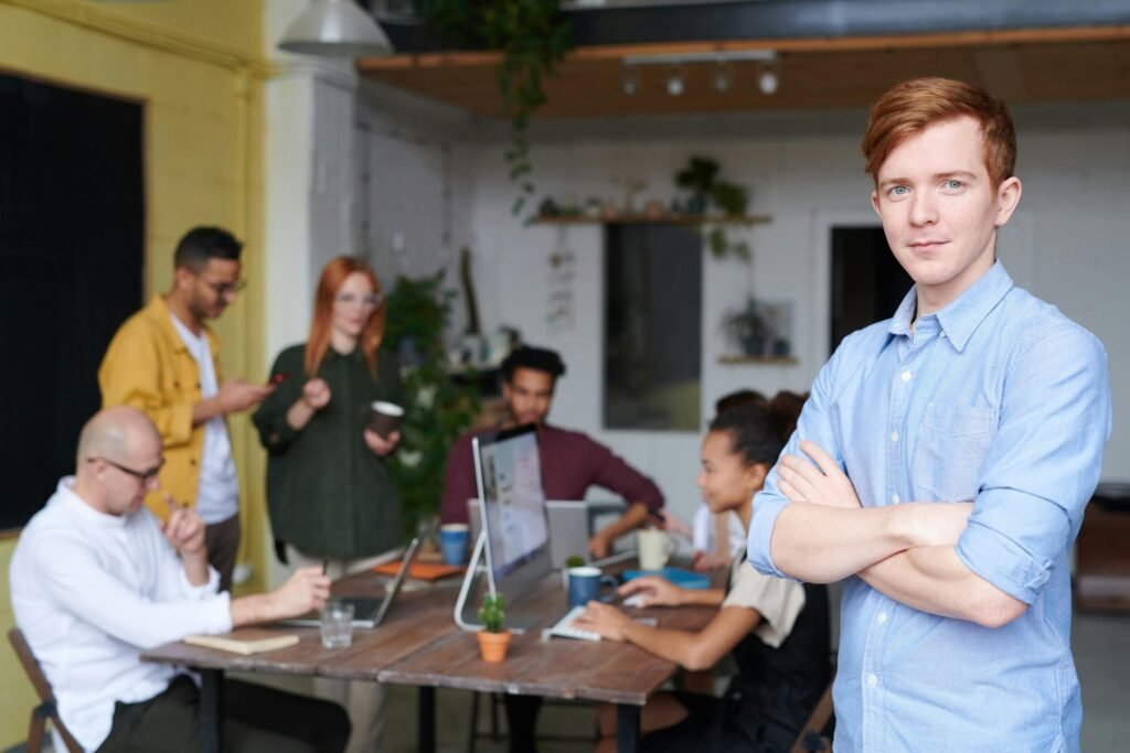 A confident young man stands in the foreground of a collaborative office environment with professionals working and engaging in conversation.